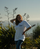 Woman in a white t-shirt standing outdoors with plants and sky in the background