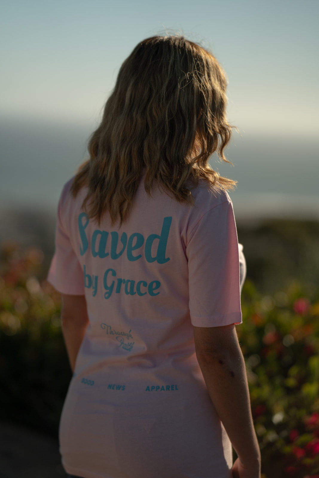 Person wearing a pink shirt with 'Saved by Grace' text, standing outdoors with blurred background