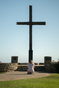 Person kneeling in front of a large wooden cross on a clear day.