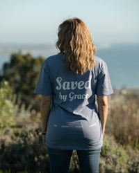 Person wearing a 'Saved by Grace' t-shirt standing on a hill with a scenic view.