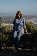 Person wearing a blue t-shirt with text, sitting on a wooden railing with a scenic background.