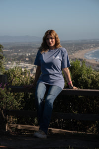 Person wearing a blue t-shirt with text, sitting on a wooden railing with a scenic background.