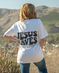 Person wearing a white t-shirt with 'Jesus Saves' on the back, standing in a scenic outdoor setting.