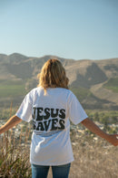 Person wearing a white t-shirt with 'Jesus Saves' on the back, standing in a scenic outdoor setting with mountains.