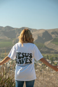 Person wearing a white t-shirt with 'Jesus Saves' on the back, standing in a scenic outdoor setting with mountains.