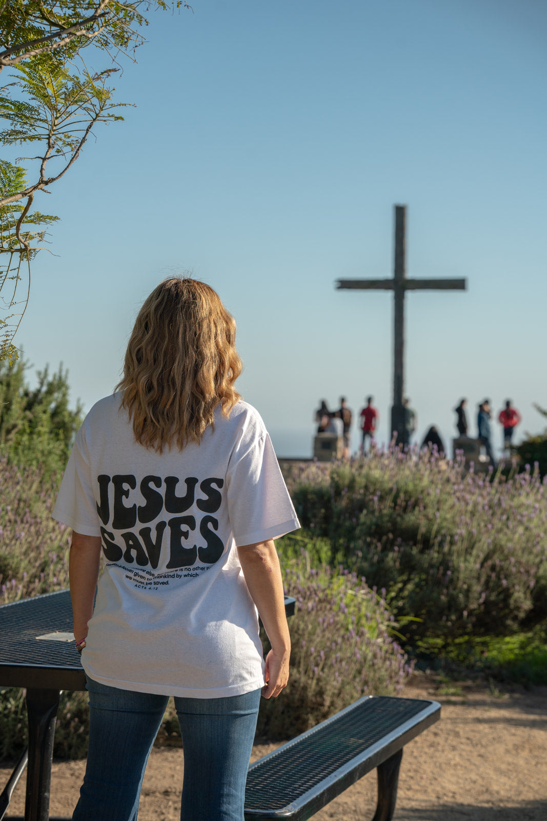 Person wearing a 'Jesus Saves' shirt standing in front of a cross with people in the background.