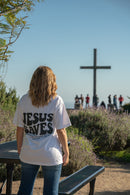 Person wearing a 'Jesus Saves' shirt standing in front of a cross with people in the background.