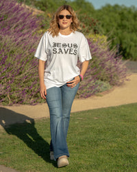 Woman wearing a 'Jesus Saves' t-shirt walking outdoors with greenery and lavender in the background