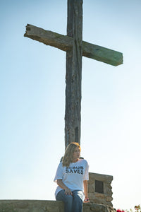 Person wearing a shirt with 'Jesus Saves' text sitting next to a wooden cross against a clear blue sky.