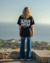 Person wearing a black t-shirt with 'Jesus Saves' text, standing on a ledge with a scenic background.