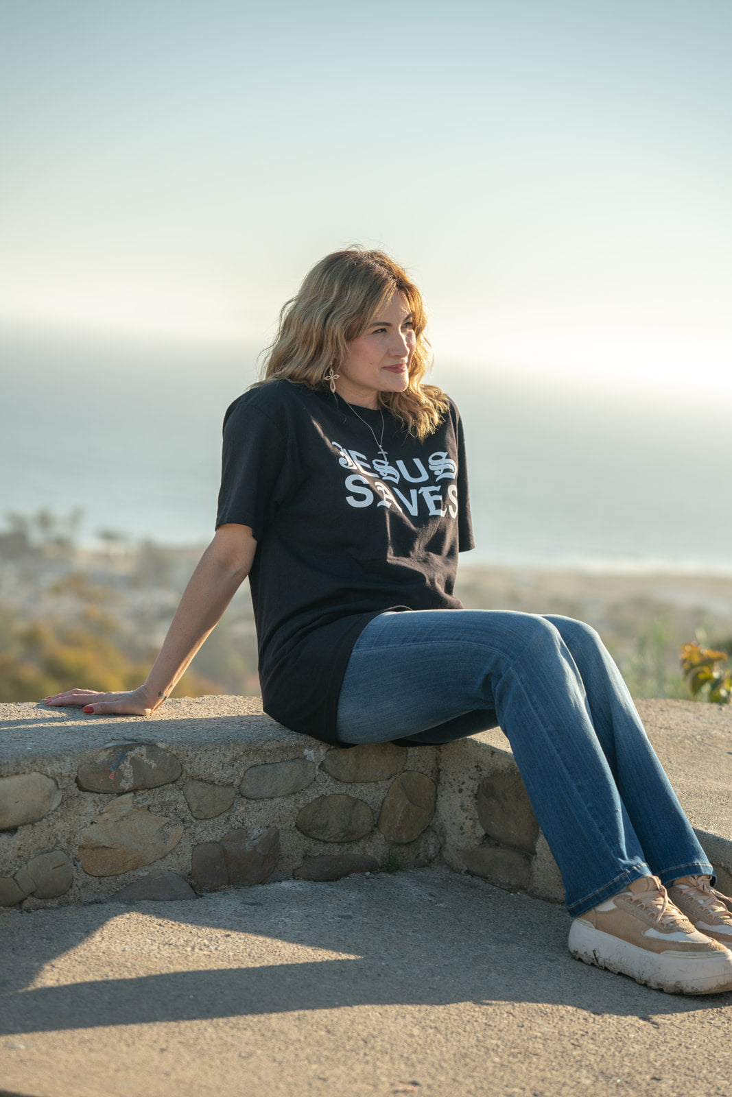 Woman wearing a black t-shirt with text, sitting on a stone wall with a scenic background.