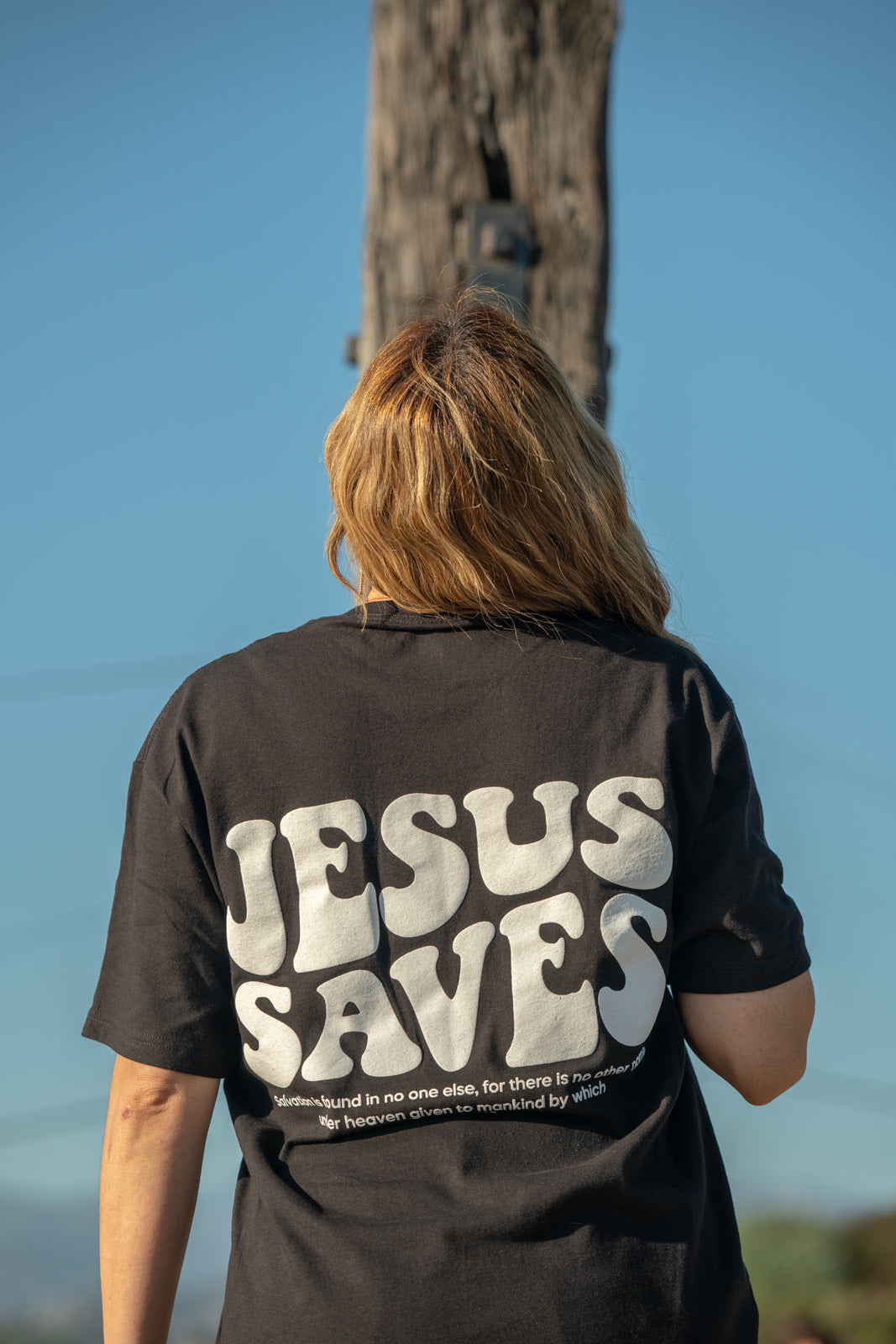Person wearing a black shirt with 'JESUS SAVES' printed on the back, standing outdoors.