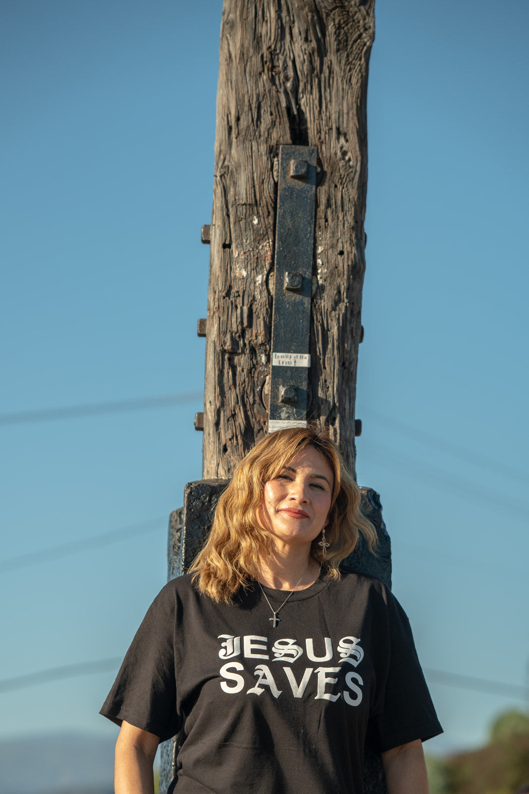Person wearing a black t-shirt with 'Jesus Saves' text standing next to a wooden post against a blue sky.