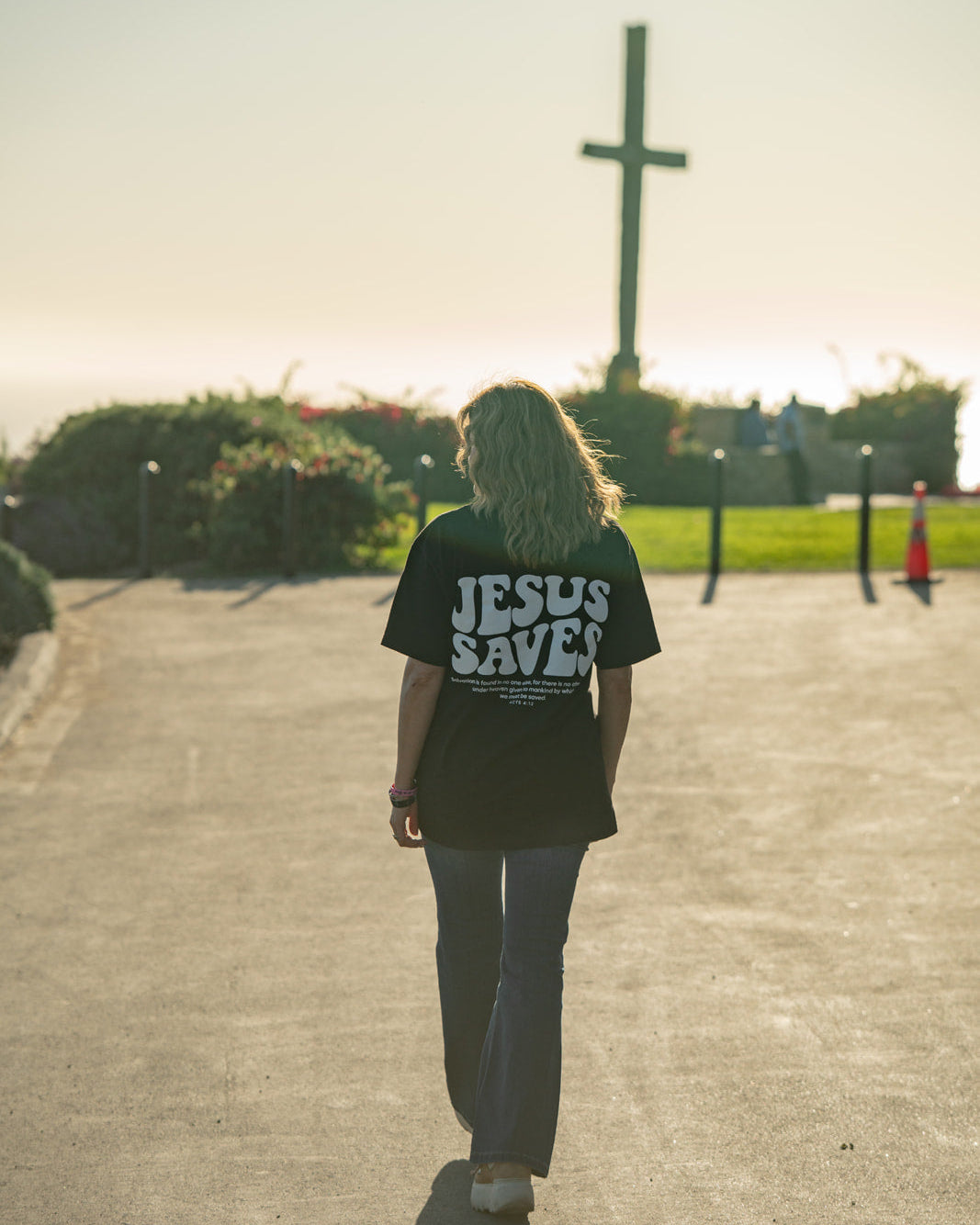Person wearing a 'Jesus Saves' shirt walking towards a cross in a park.
