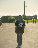 Person wearing a 'Jesus Saves' shirt walking towards a cross in a park.