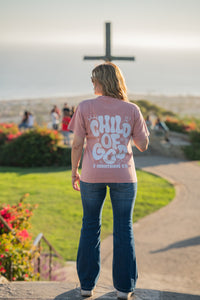 Person wearing a pink shirt with 'Child of God' text, standing on a path with a cross in the background.