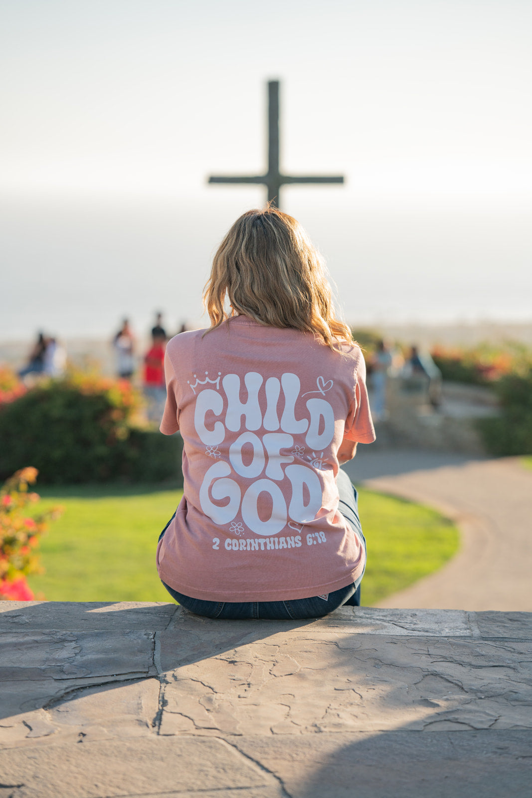 Person wearing a 'Child of God' shirt sitting on a stone wall with a cross in the background.