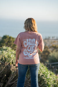 Person wearing a pink 'Child of God' shirt standing outdoors with a scenic background