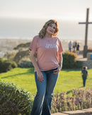 Woman wearing a pink t-shirt with text, standing outdoors with a cross and people in the background.