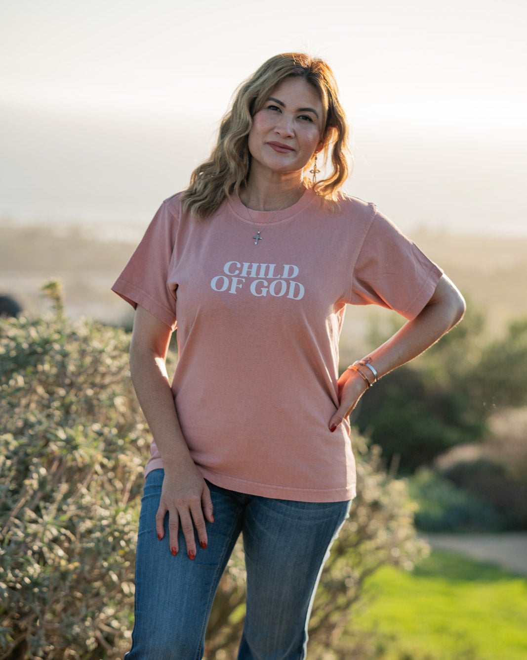 Woman wearing a 'Child of God' t-shirt outdoors with a scenic background