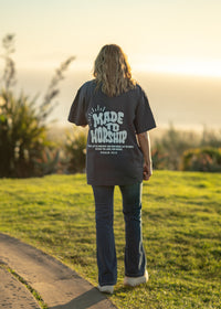 Person wearing a 'Made to Worship' shirt walking on a grassy path with a sunset in the background