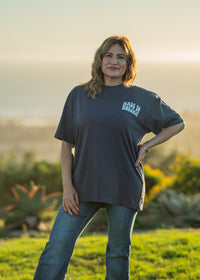 Woman wearing a dark blue t-shirt with text, standing outdoors with a scenic background.