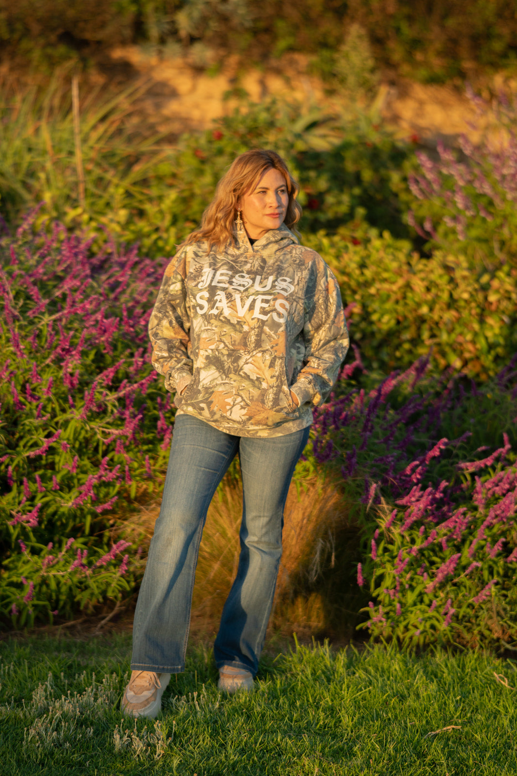 Person wearing a hoodie with 'Jesus Saves' text in a natural setting with flowers and grass.