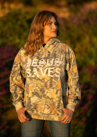 Person wearing a camouflage hoodie with 'JESUS SAVES' text in an outdoor setting