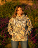 Person wearing a camouflage hoodie with 'JESUS SAVES' text, standing outdoors with greenery in the background.