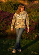 Woman wearing a camouflage hoodie with text walking through a natural setting with purple flowers.