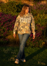 Woman wearing a camouflage hoodie with text walking through a natural setting with purple flowers.