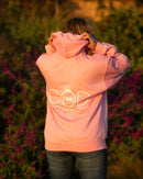 Person wearing a pink hoodie with a logo, standing in a natural setting with greenery and flowers.