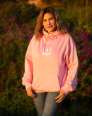 Woman wearing a pink hoodie with a logo, standing outdoors with greenery and flowers in the background.