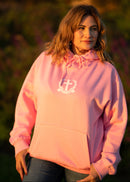 Woman wearing a pink hoodie with a logo, standing outdoors with a blurred natural background