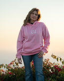 Woman wearing a pink hoodie with a logo, standing outdoors with a scenic background.