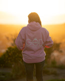 Person wearing a pink hoodie with 'GOD' printed on it, standing in a field at sunset.