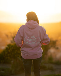 Person wearing a pink hoodie with 'GOD' printed on it, standing in a field at sunset.