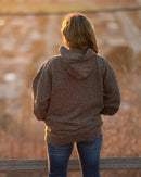 Person wearing a brown jacket and blue jeans standing by a wooden fence with a blurred natural background