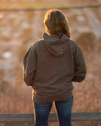 Person wearing a brown jacket and blue jeans standing by a wooden fence with a blurred natural background